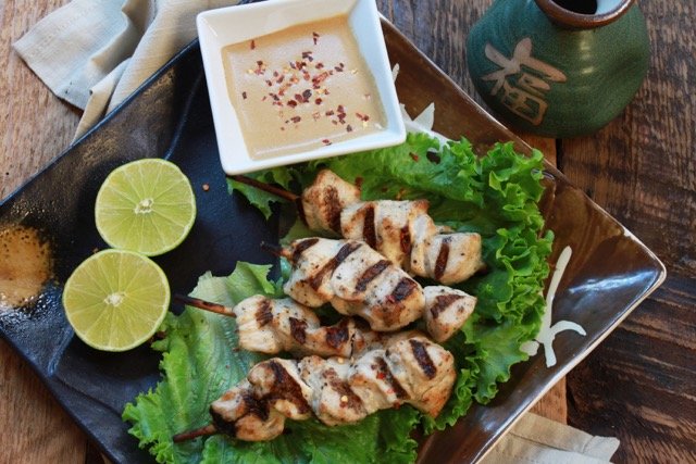 Chicken satay skewers on a bed of lettuce placed on a square brown platter with fresh limes and peanut sauce on the side.