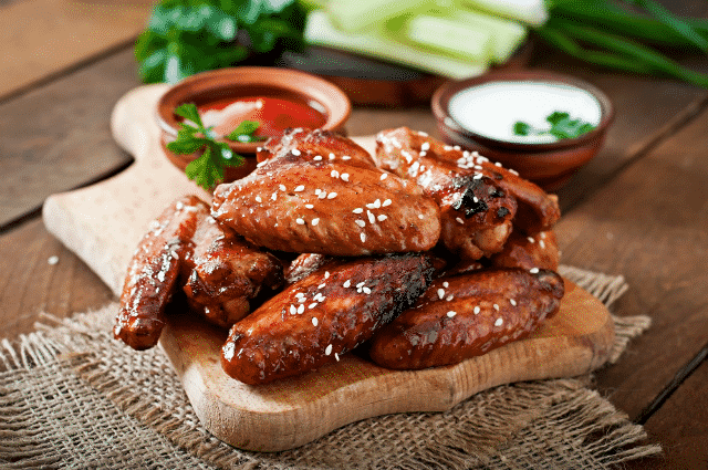 Saucy honey garlic chicken wings piled on top of a wooden cutting board with sauces and celery on the side, on top of a burlap placemat.