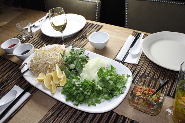 a table set with plates and green herb toppings for Vietnamese pho ga on a white serving platter 