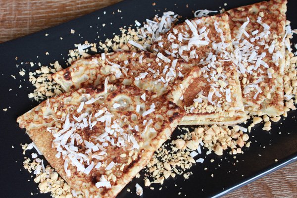 Nutella and Coconut Pancake crepes sprinkled with coconut flakes and garnished with chopped peanuts on a black plate on top of .a wooden board
