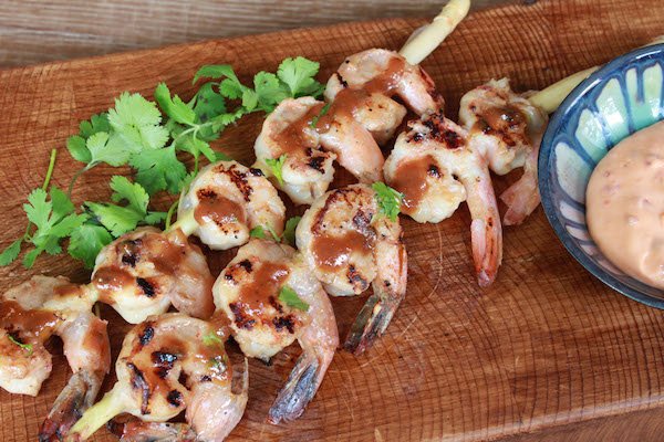 Grilled shrimp skewers on a brown platter with dipping sauce in a blue and white bowl and cilantro garnish.