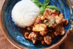 Tender pieces of chicken with chili jam on a blue and brown bowl with a side of white rice on the side and a sprig of fresh basil..