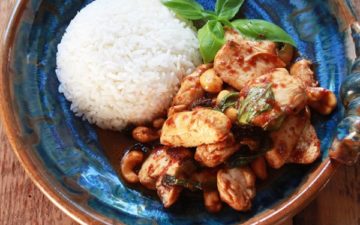 Tender pieces of chicken with chili jam on a blue and brown bowl with a side of white rice on the side and a sprig of fresh basil..