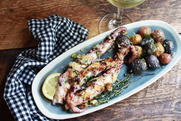 Grilled octopus topped with herbs on a long serving platter with roasted baby potatoes, a checkered napkin and glass of wine on the side, on top of a wooden board.