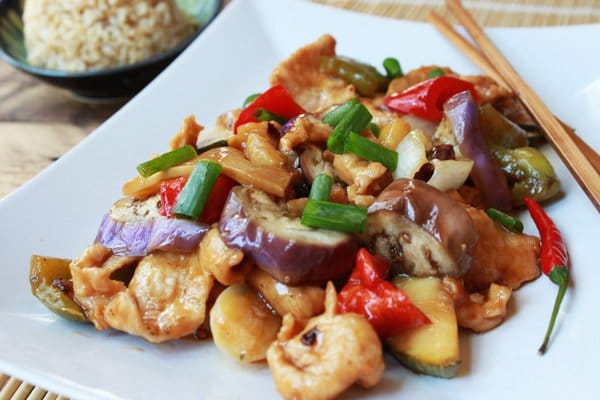Szechuan chicken and eggplant on a white plate with chopsticks and a bowl of brown rice on the side.