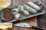 Fresh Vietnamese shrimp spring rolls on a long gray platter with a peanut sauce on the side on top of a wooden board with a knife and napkin.