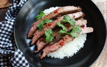 Slices of Grilled Asian Flank Steak on a bed of rice in a black bowl, on a wood table with a gingham napkin.