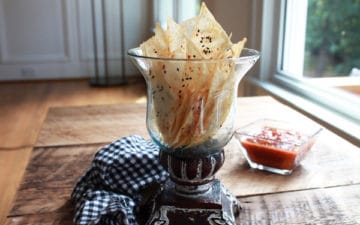 A clear bowl filled with seasoned baked wonton chips on top of a wooden board with a checkered napkin and salsa on the side.