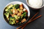 a bowl of stir-fry broccoli and cubed tofu with chopsticks and a bowl of white rice on the side