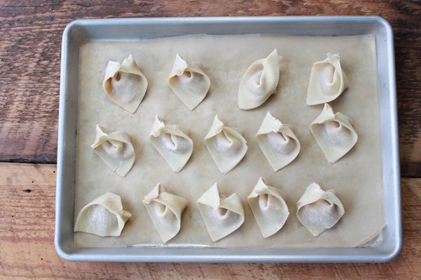 Rows of uncooked wontons placed on a baking sheet on top of a wooden board.