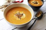 A white bowl filled with pumpkin coconut soup, with a spoons and red chilis on the side.