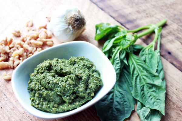 A small white bowl filled with a pesto sauce with fresh basil leaves on the side and walnuts in the background on top of a wooden board.
