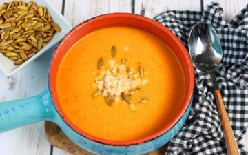 Sweet and Spicy Chilled Carrot Soup in a blue bowl garnished with pumpkin seeds on a white board with a spoon and gingham napkin beside the bowl