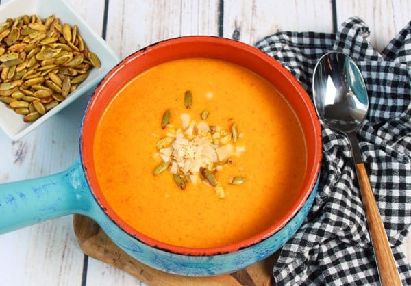 Sweet and Spicy Chilled Carrot Soup in a blue bowl garnished with pumpkin seeds on a white board with a spoon and gingham napkin beside the bowl