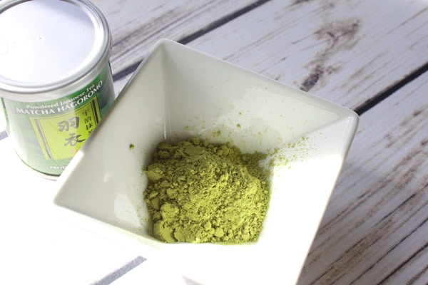 Matcha green tea powder in a white bowl with a can of matcha powder beside it on a white plank wooden board.