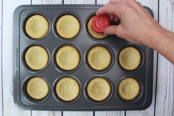 Baked sugar cookie cups in a muffin tin with a red capped spice jar used to place an indentation in the cookie cups on a white plank wooden board.