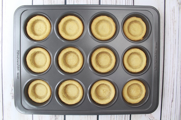 Baked sugar cookie cups with indentations in a muffin tin on a white plank wooden board.