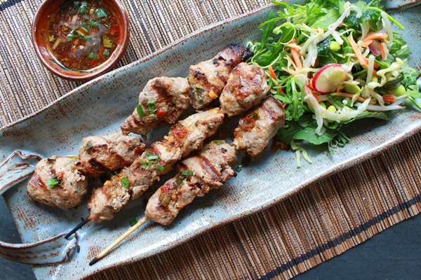 A blue platter with three skewers of Vietnamese pork tenderloins with a side of crispy salad and sauce on the side.