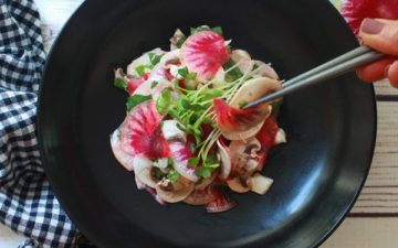 A vibrant mushroom and radish salad in a black bowl with sprouts on top and silver chopsticks on the side.