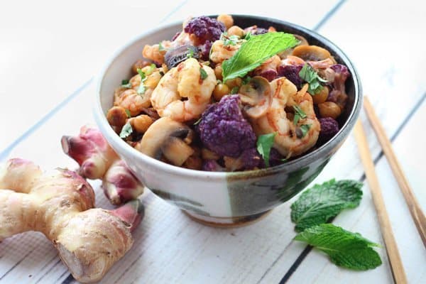 A black and white bowl filled with shrimp sambal and roasted cauliflower and chickpeas topped with a fresh mint leaf with ginger bulbs on the side on top of a wooden white board.