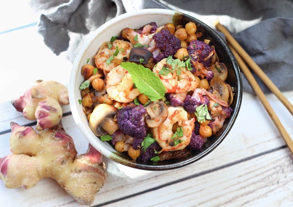Shrimp sambal with cauliflower chickpeas and ginger in a black and white bowl with chopsticks and fresh ginger and a grey napkin on the side on a white plank wooden board.
