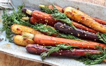 roasted rainbow carrots with tops on placed on a white and beige oval plate on a wooden board