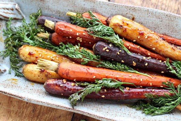 Roasted rainbow carrots with green tops on placed on a white and beige oval plate garnished with sesame seeds on top of a wooden board.