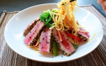 Sesame crusted tuna in a white bowl with Asian noodles help up by chopsticks on a bamboo placemat.
