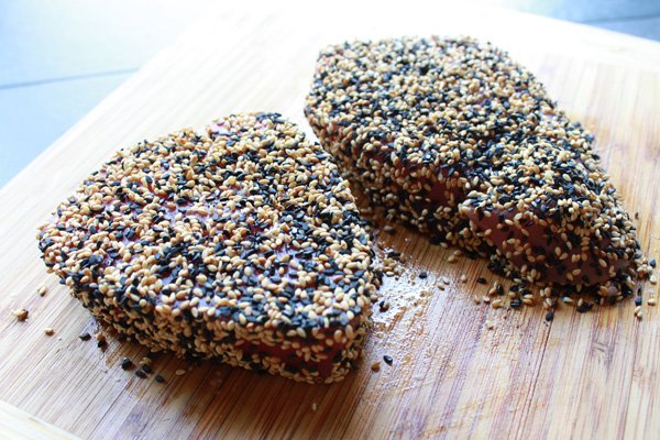 Two tuna steaks crusted with a mixture of black and white sesame seeds on a wooden cutting board.