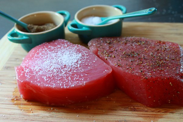 Raw yellowfin tuna steaks sprinkled with salt and pepper on a cutting board with salt and pepper pots on the side.