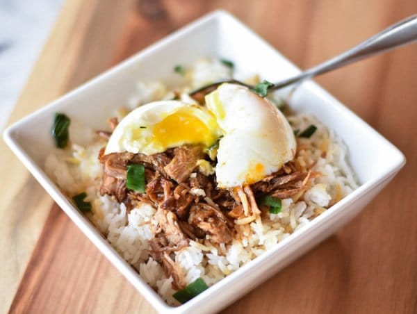 Instant Pot Korean Inspired Pulled Pork over rice in a white square bowl topped with chopped green onions and an runny egg with a fork in the bowl on a wooden cutting board.