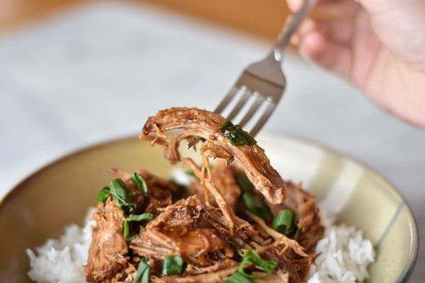 Korean Instant Pot Korean Inspired Pulled Pork on a bed of rice garnished with sliced green onions in a beige bowl with someone holding a fork with pulled pork on it.