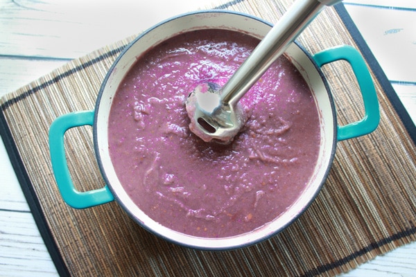 Purple potato soup in a blue and white pot with an immersion blender inserted inside on top of a bamboo placemat placed on a white plank wooden board.