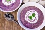 Purple potato and cauliflower soup in white bowls with a topping of sour cream and garnished with sprigs of cilantro with a spoon and beige napkin on the side on a bamboo placemat
