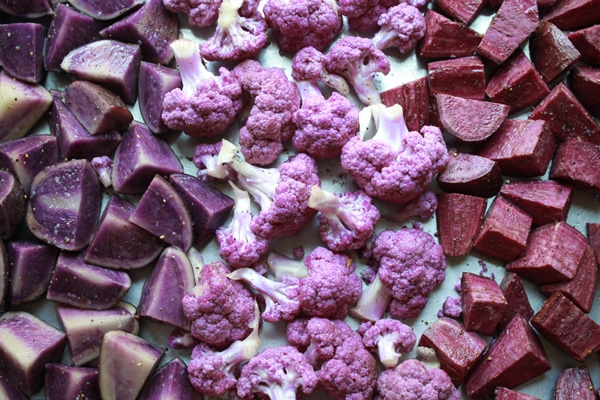 Purple cauliflower florets and cubed purple potatoes on a baking sheet ready to roast in the oven.