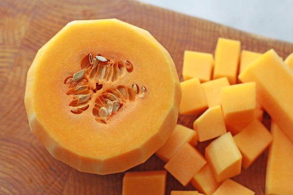 A peeled and halved butternut squash with seeds and cubed butternut squash pieces on the side on top of a wooden cutting board.