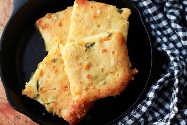 Squares of jalapeño cornbread in a small cast iron skillet on top of a wooden board with a checkered napkin on the side.