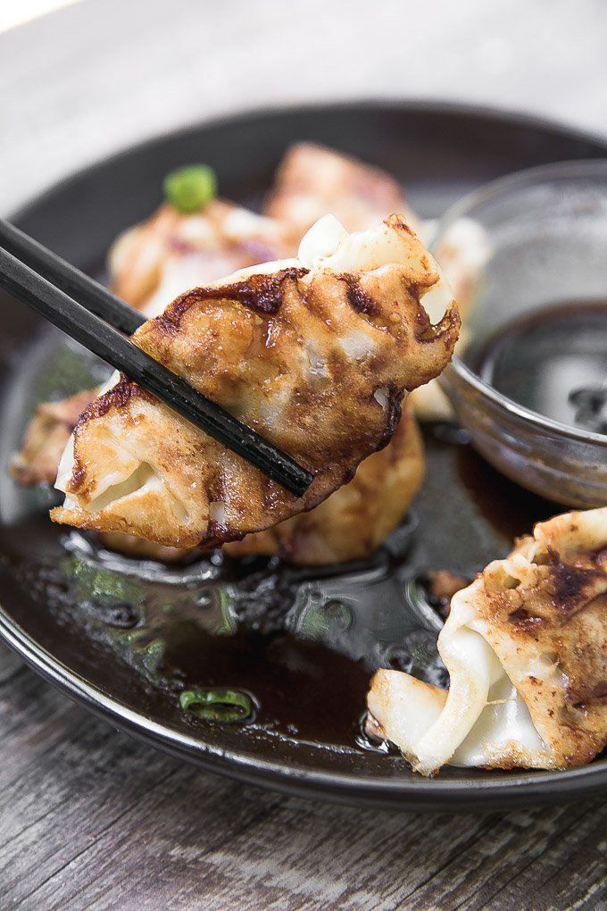 Pork potstickers held by chopsticks over a black plate with a clear glass bowl of dipping sauce on the side on top of a dark wooden board.