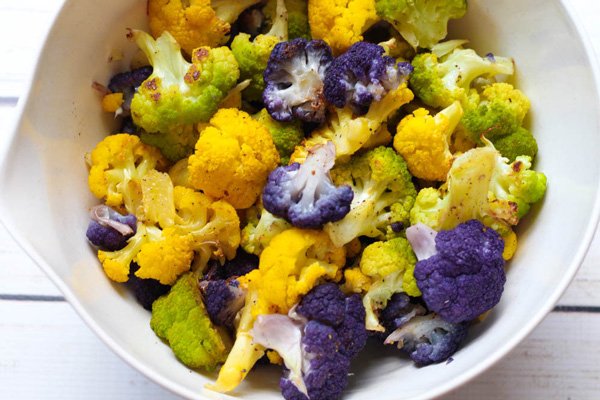 Roasted purple, green, and orange cauliflower florets in a white bowl on top of a white plank wooden board.