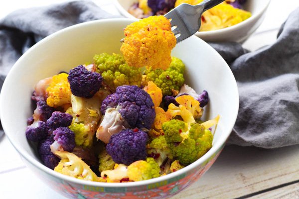 Tri-colored cauliflower florets in a white bowl with an orange piece of cauliflower held on a fork with a grey napkin on the side on top of a wooden board.