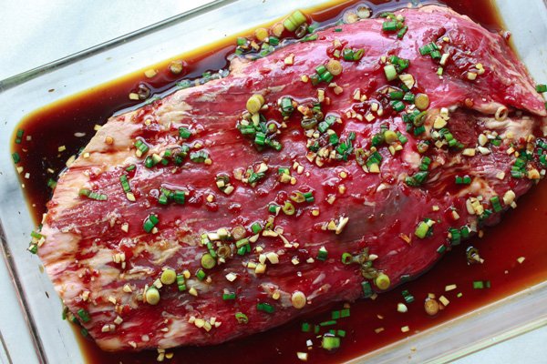Raw flank steak marinating in a clear glass pan.