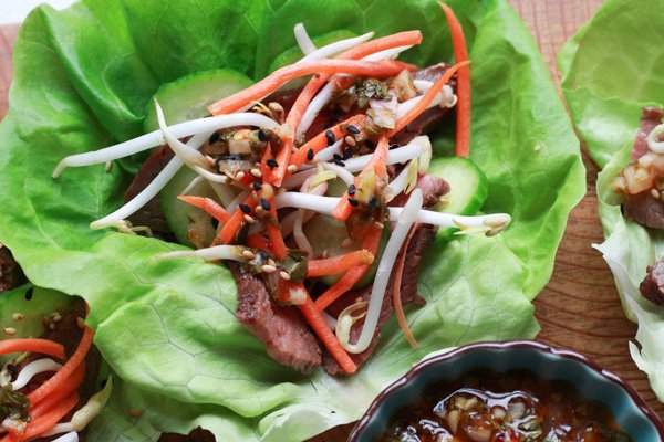 Grilled flank steak lettuce cups with a side of nuoc cham sauce in a dark blue bowl on top of a wooden board.