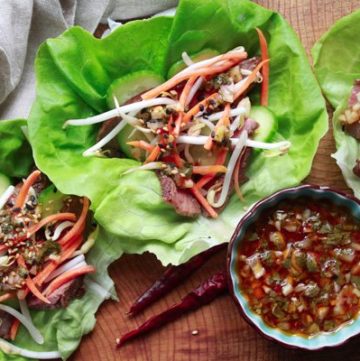 Overhead view of grilled flank steak lettuce cups on a wooden board with a side of nuoc cham sauce in a dark blue bowl with hot peppers.