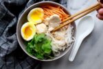 miso chicken noodle soup with noodles, cilantro, hard boiled egg, carrots and chicken in a bowl with chicken being held with chopsticks and a soup spoon on the side and a dark grey checked napkin on the side on a white marble board