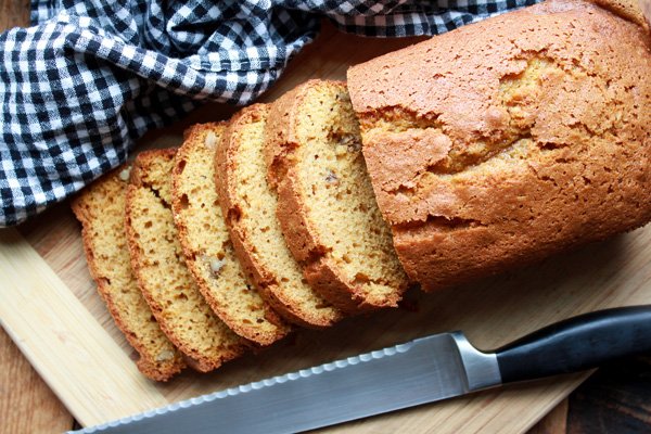 A loaf of pumpkin bread partially sliced on a wooden cutting board with  a bread knife and checkered napkin on the side.