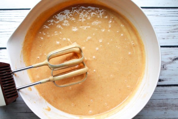 A white mixing bowl filled with pumpkin bread batter with mixing beaters on the side on top of a white wooden plank board.