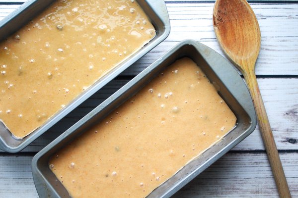 Two aluminum bread loaf pans filled with pumpkin bread batter with a wooden spoon on the side on top of a white wooden plank board.