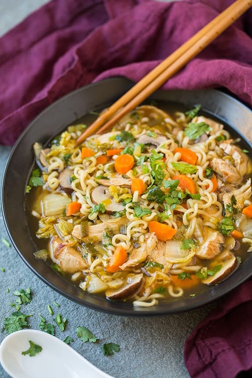 A large black bowl filled with Asian chicken noodle soup with ramel noodles and veggies with a pair of chopsticks and a white spoon on the side.