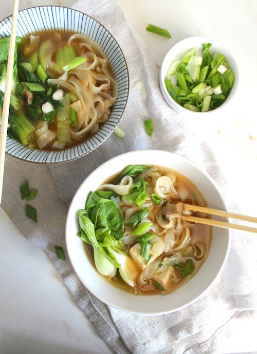 A white bowl filled with Asian vegetable noodle soup with baby bok choy and tofu with a pair of chopsticks inserted on top of a white linen napkin.