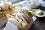 Fluffy steamed bao buns filled with eggs and avocado slices topped with fresh bean sprouts sitting in a taco holder with a small bowl of bean sprouts in the background.